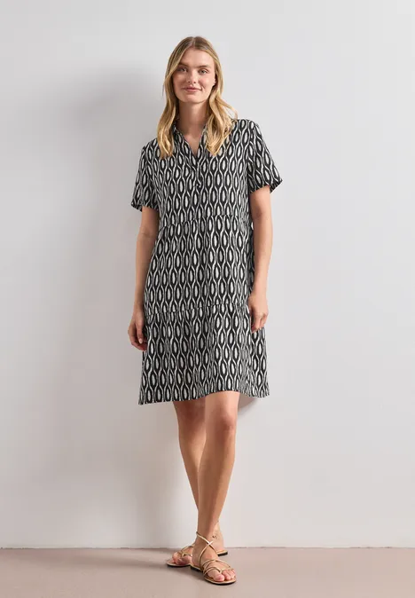 Frau in knielangem, schwarz-weißgemustertem Kleid mit kurzen Ärmeln und Sandalen vor hellem Hintergrund.
