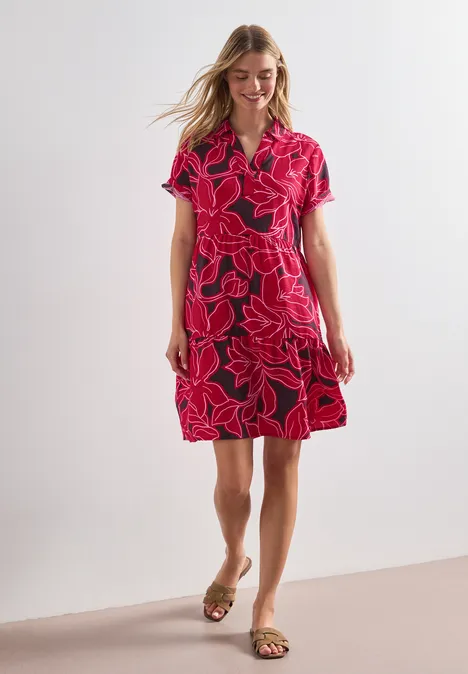 Frau in knielangem rotem Kleid mit großem rosa Blumenmuster, kurzen Ärmeln und Sandalen vor hellem Hintergrund.