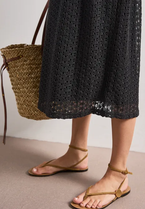 Frau in schwarzem Häkelkleid mit Basttasche und flachen, olivgrünen, geschnürten Sandalen.