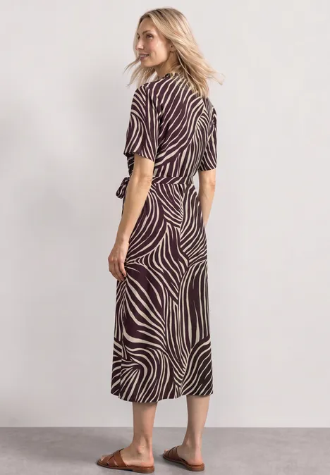 Frau im midi-Kleid mit Zebramuster, kurzen Ärmeln und Gürtel, trägt flache Sandalen vor neutralem Hintergrund.