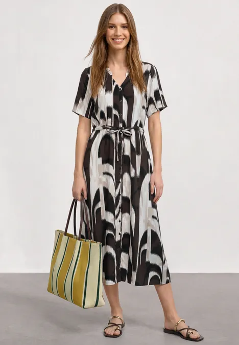 Frau im knieumspielenden Schwarz-Weiß-Kleid mit Taillengürtel, trägt schwarze Sandalen und grüne Streifen-Tasche.