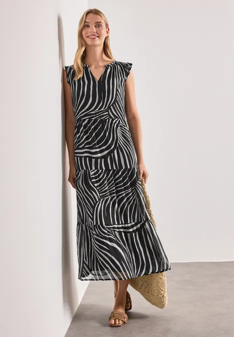Femme appuyée à un mur dans une robe mi-longue zébrée noire et blanche, ceinture et sac en paille.