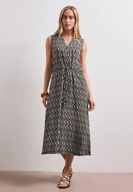 Frau in ärmellosem, gemustertem Maxikleid mit Bindegürtel und Sandalen.