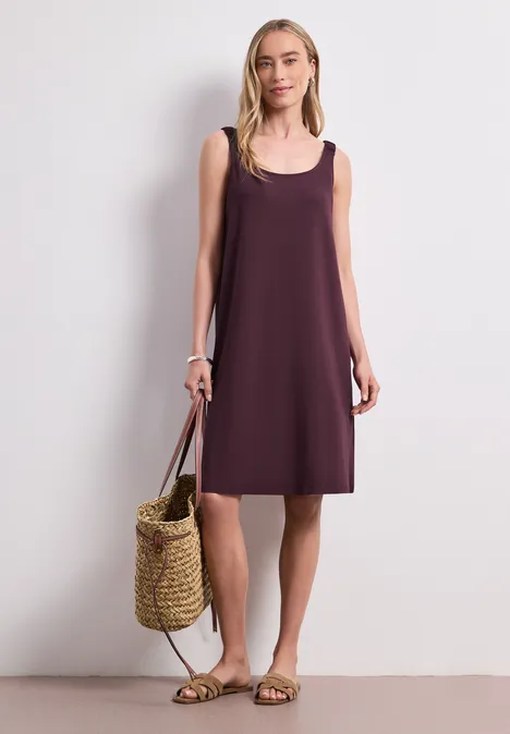 Frau trägt ärmelloses, locker sitzendes lila Kleid, hält Strohtasche und Sandalen.