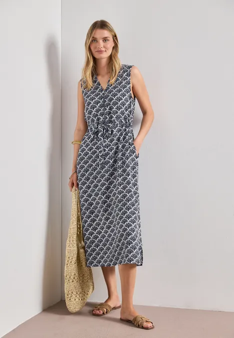 Frau trägt ärmelloses, gemustertes Midikleid in Blau-Weiß mit V-Ausschnitt, gebundener Taille, Basttasche und Sandalen.