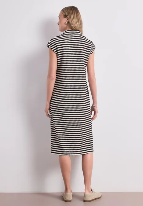 Femme de dos devant un mur blanc, portant une robe midi rayée noir et blanc et des chaussures beiges.