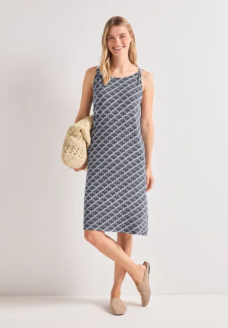 Frau in ärmellosem, kariertem Midikleid mit maritimem Muster, trägt Basttasche und bequeme Espadrilles.
