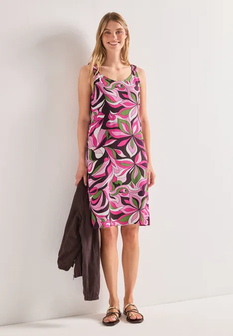 Frau in knielangem, pink-grün gemustertem Kleid, Sandalen tragend, mit Jacke am Arm vor heller Wand.
