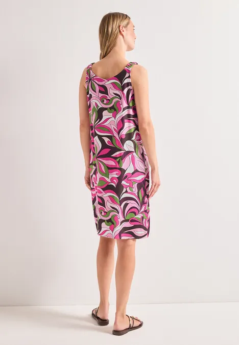 Frau von hinten in ärmellosem Midikleid mit pink-weiß-schwarzem Muster und Sandalen vor hellem Hintergrund.