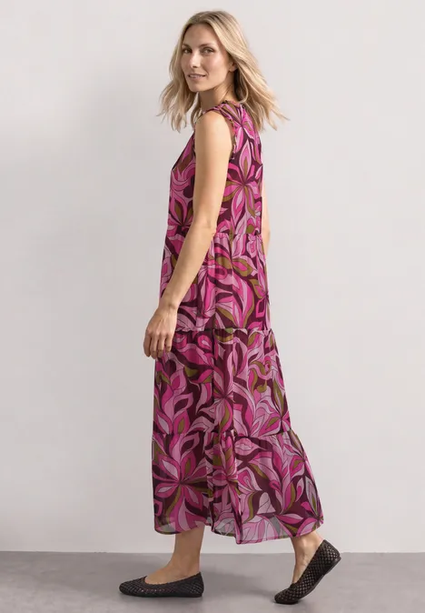 Frau im seitlichen Profil trägt langes ärmelloses Maxikleid mit pink-lila Blumenmuster und flachen Schuhen.