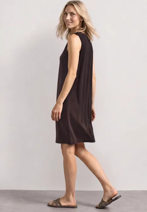 Femme en robe noire sans manches et sandales marchant de profil sur fond clair.