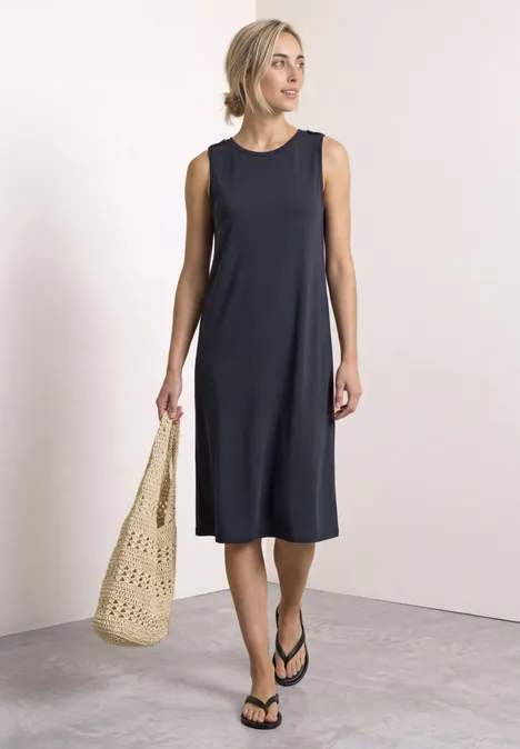 Vrouw in mouwloze zwarte midijurk met strooien tas en sandalen tegen neutrale achtergrond.