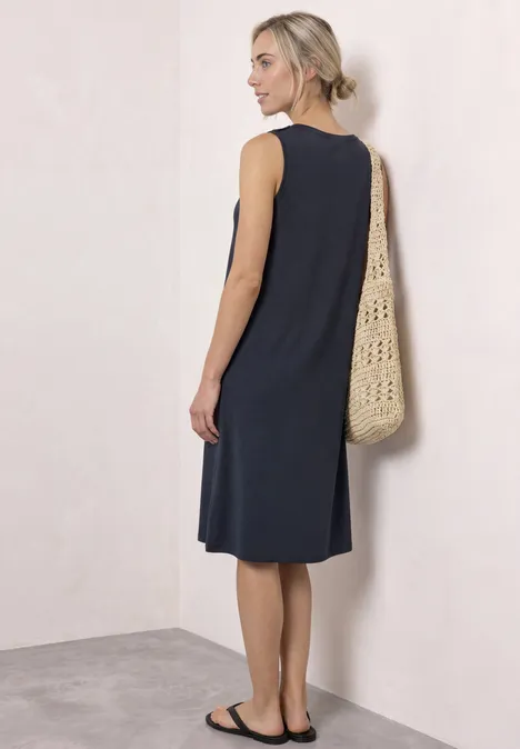Vrouw in mouwloze donkerblauwe midijurk, met gevlochten tas over de schouder, staand voor een lichte muur.