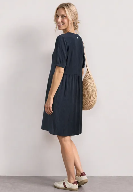 Knielanges Kleid mit Schleifendetail darkest blue Frau in lockerem dunkelblauem Kleid mit kurzen Ärmeln trägt eine Flecht-Tasche und Slip-on-Schuhe, seitlich stehend.