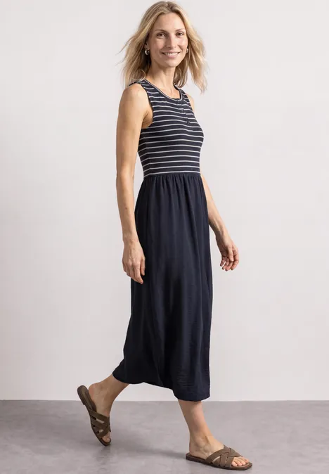 Eine lächelnde Frau in einem ärmellosen, marineblau-gestreiften Top und Midirock mit Sandalen.