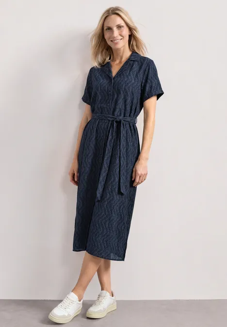 Personne en robe midi bleu marine à ceinture et baskets blanches devant un fond clair.