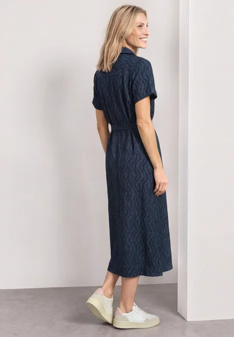 Une femme porte une robe-chemise midi bleu marine à manches courtes et des baskets blanches.