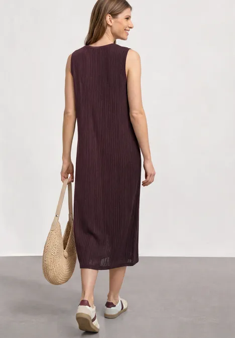 Vrouw van achter in een mouwloze midi jurk bordeaux, met rieten tas en witte plateau sneakers.