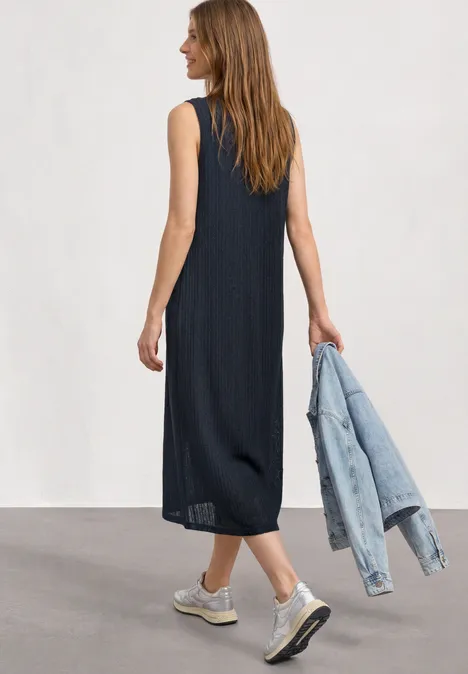 Vrouw van achteren in zwarte mouwloze midijurk, draagt een spijkerjasje en witte sneakers.