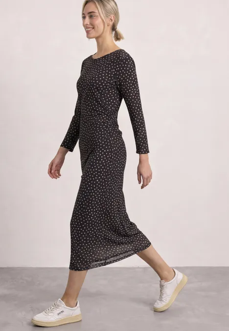 Een glimlachende vrouw loopt in een zwarte midi-jurk met witte stippen en beige sneakers.