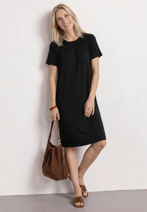 Robe en jersey à longueur de genou et col rond Black Femme en robe noire simple jusqu’aux genoux, sac marron et sandales, posant devant un mur blanc