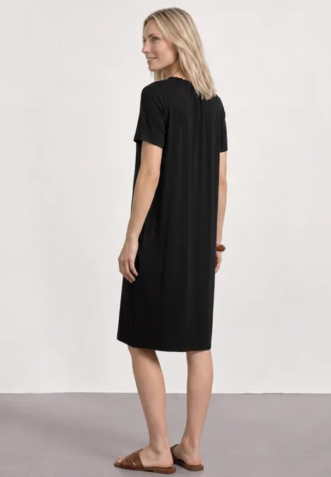 Robe en jersey à longueur de genou et col rond Black Femme aux cheveux blonds portant une robe midi noire et des sandales, de dos devant un mur blanc.