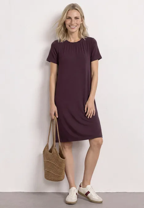Eine Frau trägt ein schlichtes, knielanges, violettes T-Shirt-Kleid, hält eine geflochtene Strohtasche und weiße Sneaker.