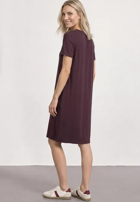 Eine Frau steht seitlich, trägt ein lockeres lila T-Shirt-Kleid und sportliche weiße Sneakers.
