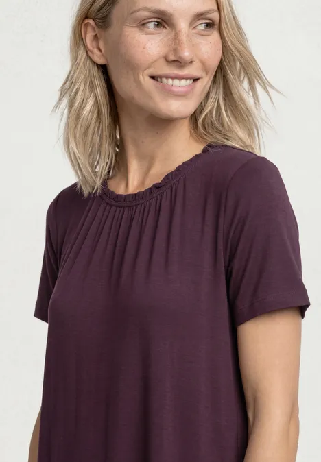 Frau mit schulterlangem blonden Haar trägt violettes locker sitzendes T-Shirt vor hellem Hintergrund