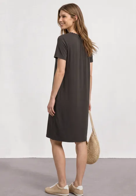 Frau von hinten in lockerem, knielangem, dunkelgrauem Kleid mit Sneakers und Strohtasche, helles Studio-Setting.