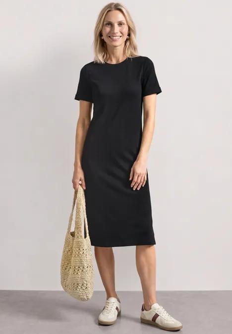 Midikleid in Rippstruktur mit Rundhals Black Frau in schlichtem schwarzem T-Shirt-Kleid, weiße Sneaker und Netztasche, vor schlichtem Hintergrund.
