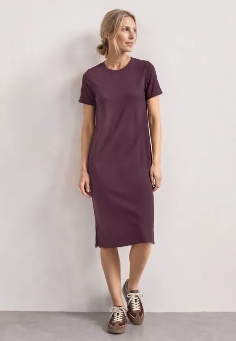 Vrouw in eenvoudig paarse midi T-shirtjurk met witte sneakers voor een neutrale achtergrond.