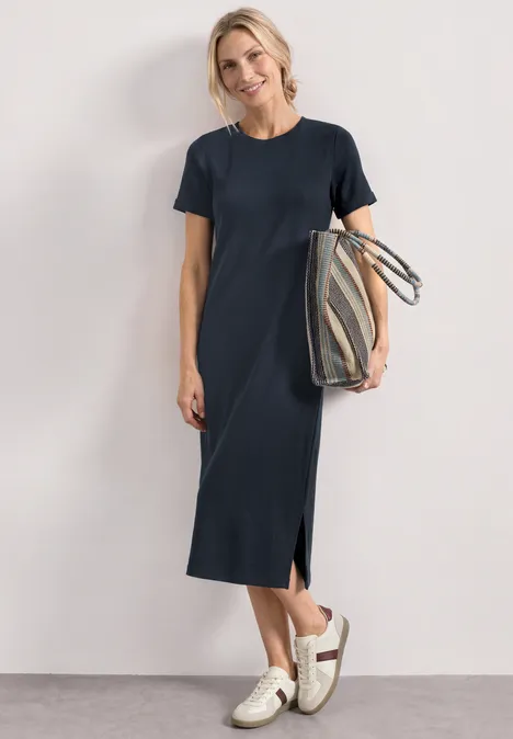 Frau in schlichtem navy Midi-Kleid mit Seitenschlitz und Taschen, kombiniert mit weißen Sneakern und Streifentasche.