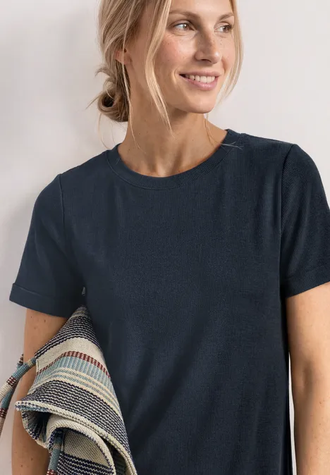 Frau mit dunkelblauem T-Shirt, seitliche Pose, hält gemusterten Stoff unter dem Arm.