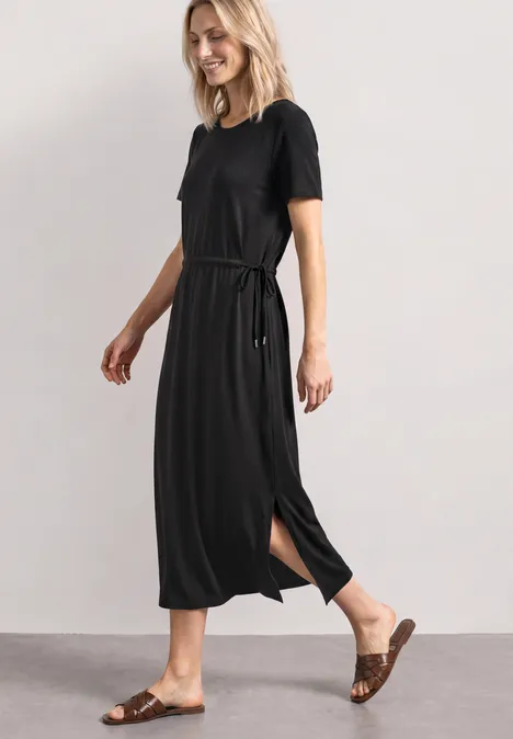 Une femme porte une robe midi noire à manches courtes, ceinture et fente sur le côté, associée à des sandales marron.