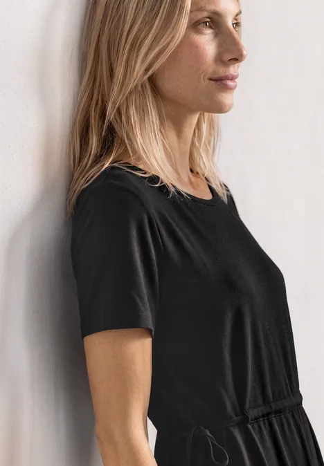 Femme de profil en robe noire à manches courtes adossée à un mur, regard tourné vers la droite.