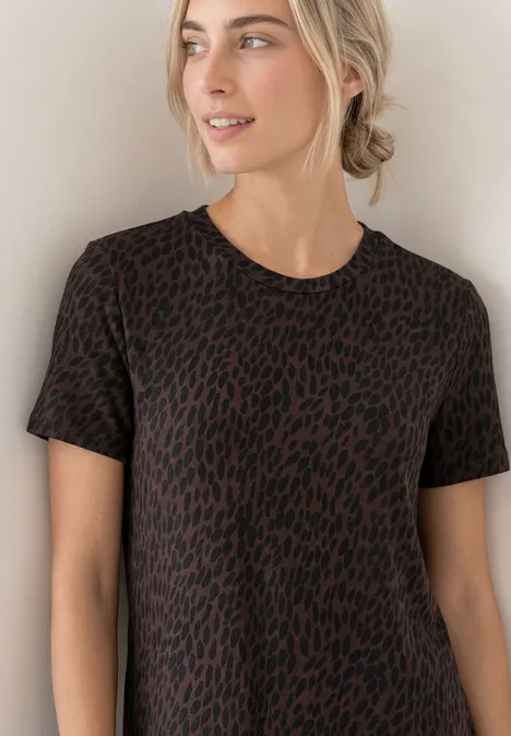 Frau mit blond zurückgebundenem Haar trägt kurzärmliges T-Shirt mit braun-schwarzem Leopardenmuster vor hellem Hintergrund.