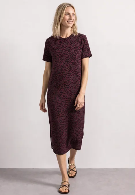 Eine lächelnde Frau im kniehohen, kurzärmeligen, rot-schwarzen Kleid mit schwarzen Sandalen vor weißem Hintergrund.