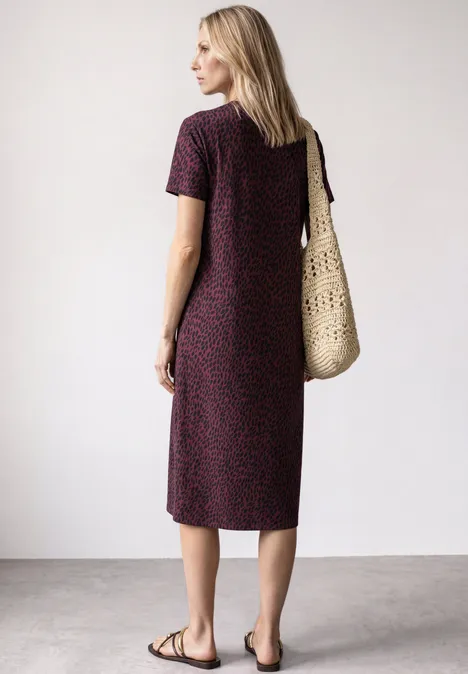 Frau im gemusterten Midi-Kleid, trägt Basttasche und flache Sandalen, steht seitlich vor hellem Hintergrund.