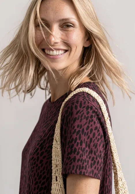 Blonde Frau in burgunderrotem Top mit Leopardenmuster und geflochtener Strohtasche lächelt.