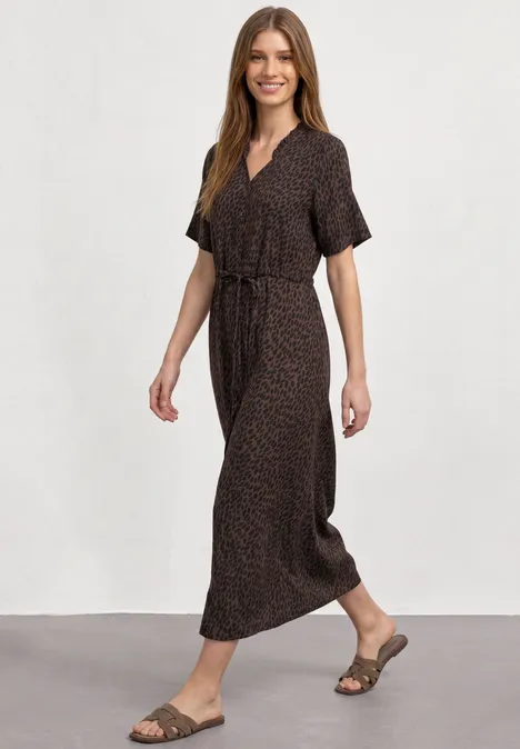 Femme en robe longue marron à manches courtes, ceinture et sandales, marchant en souriant.