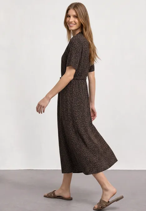 Femme en robe midi marron à motifs et sandales, souriante, se tourne en arrière.
