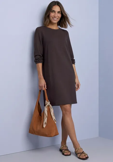 Frau in lockerem, braunem Kleid mit lässiger Handtasche und Sandalen vor pastellfarbener Wand.
