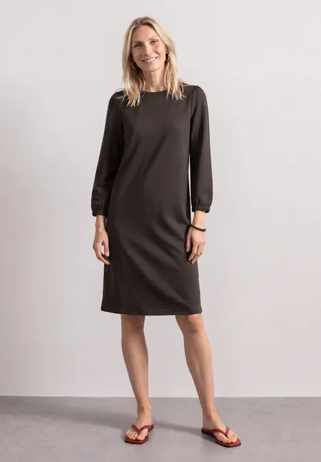 Vrouw in zwarte, knielange jurk met lange mouwen en teenslippers voor een lichte achtergrond.