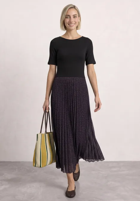 Een vrouw draagt een zwarte top, een geplisseerde paarse midirok, zwarte ballerina’s en een geel-wit gestreepte tas.