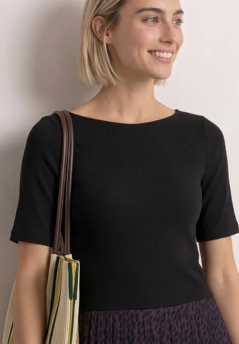 Een glimlachende vrouw in zwart shirt en patroonrok met schoudertas, leunend tegen een muur.