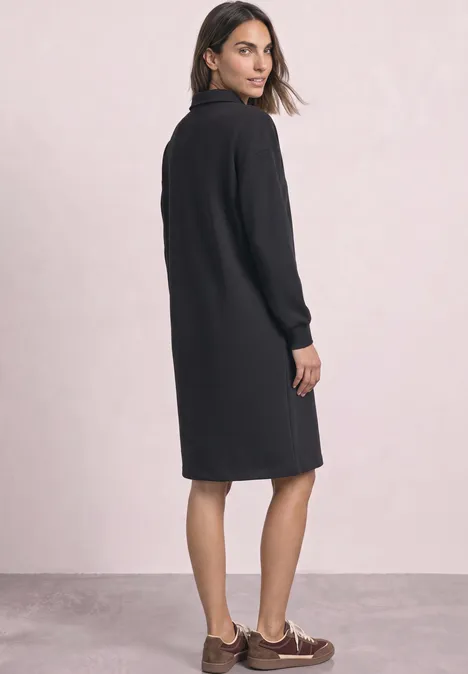 Frau in schwarzem, knielangem Kleid mit langen Ärmeln, seitlich stehend und über die Schulter blickend.