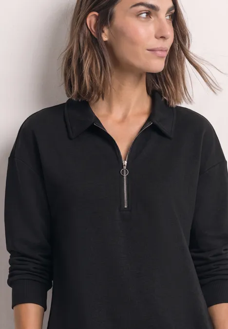 Frau in schwarzem Langarmshirt mit Reißverschluss und Kragen, schulterlanges braunes Haar, frontalaufnahme.