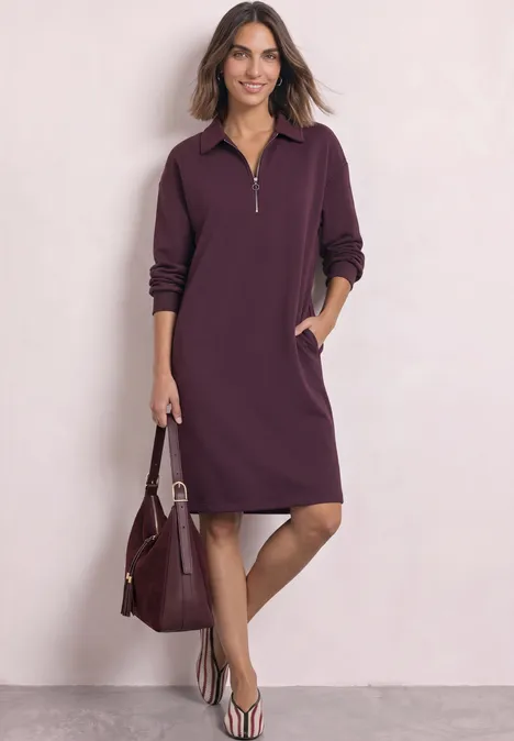 Femme en robe violette manches longues à demi-fermeture éclair et poches, avec sac assorti et sandales.