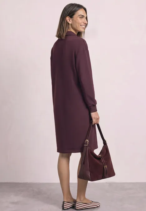 Une femme porte une robe violette tombant aux genoux, ample, et tient un sac assorti.